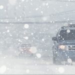 A photo of a blue SUV driving on the road in a winter snowstorm.